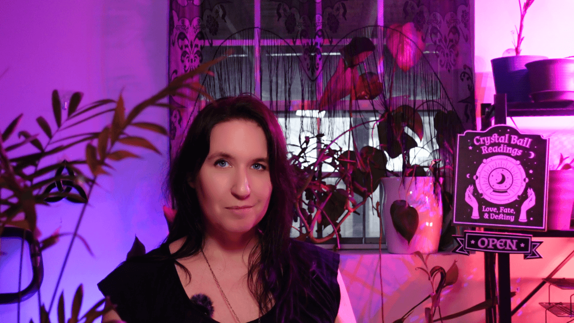 Woman sitting in a dimly lit room with a neon "open" sign and a "crystal ball readings" sign, surrounded by plants.